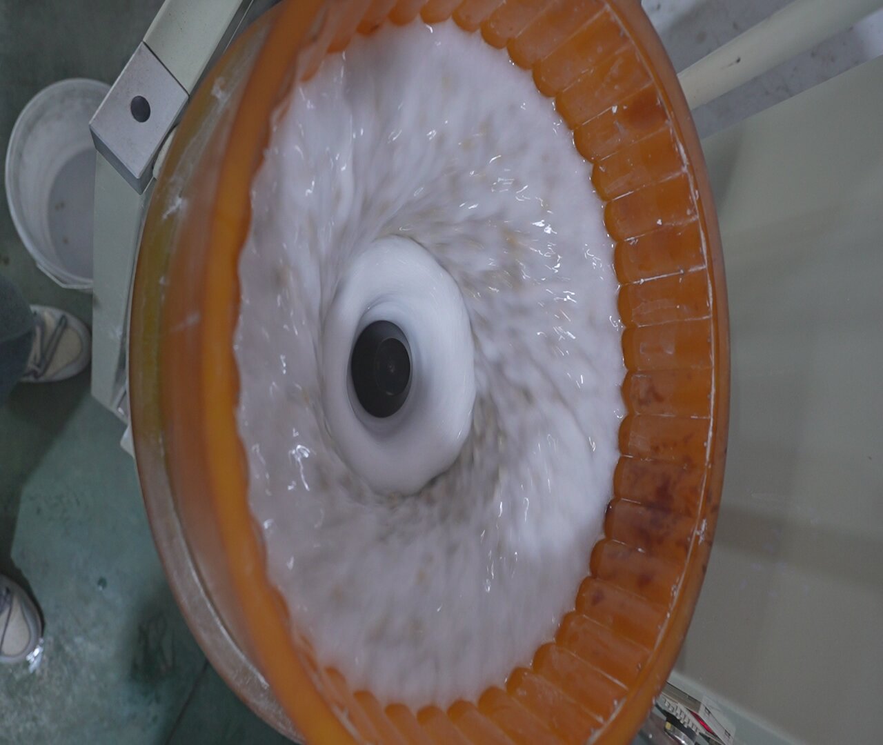 Bên trong máy bát đang chạy: lớp lót polyurethane màu cam, nước và hạt mài chuyển động theo dòng xoắn ốc quanh trục giữa