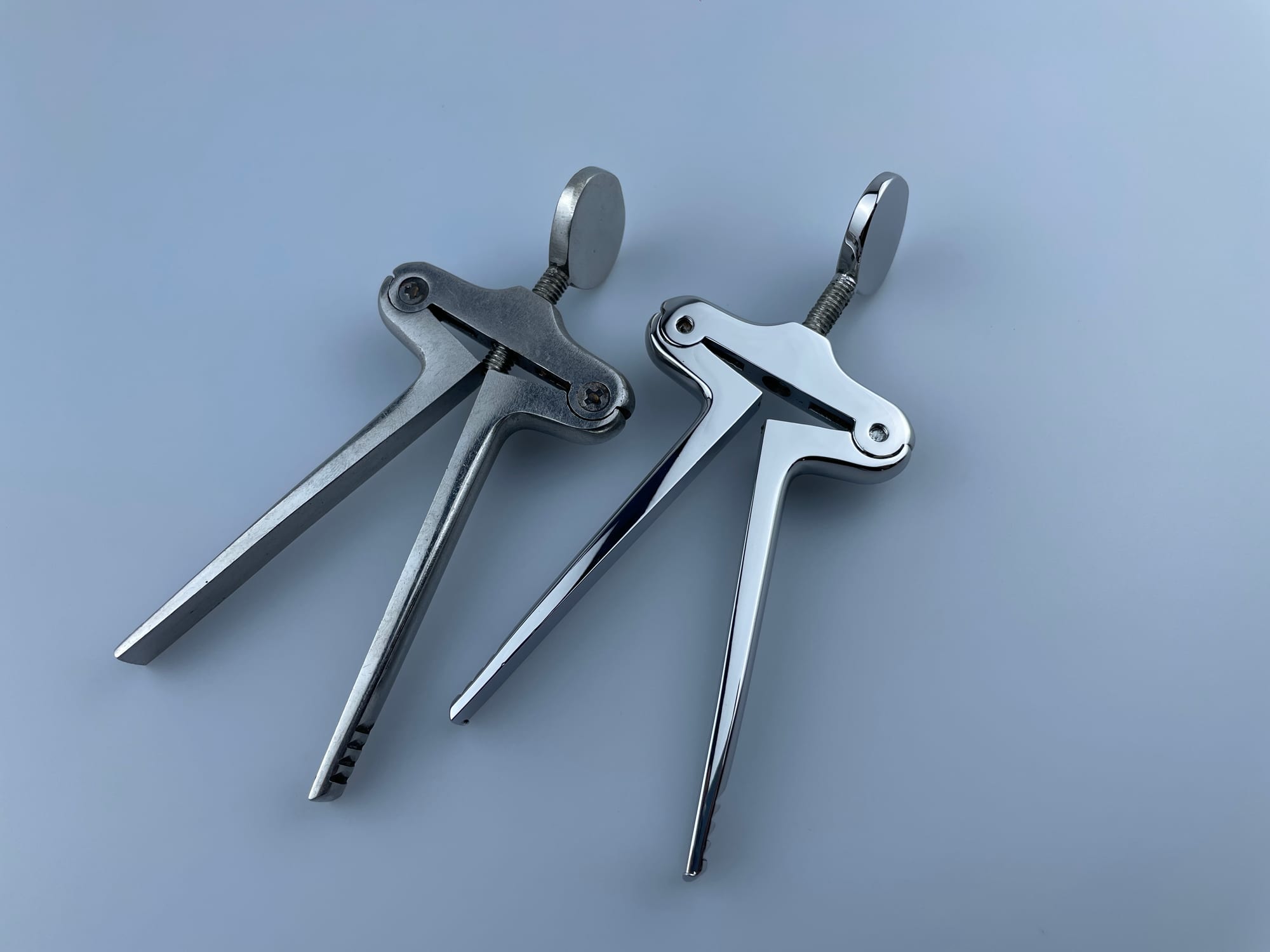 The image shows a pair of stainless steel pliers sitting on top of a blue surface. The pliers have a sleek, modern design with a silver finish. They are connected to each other by a thin metal rod, an